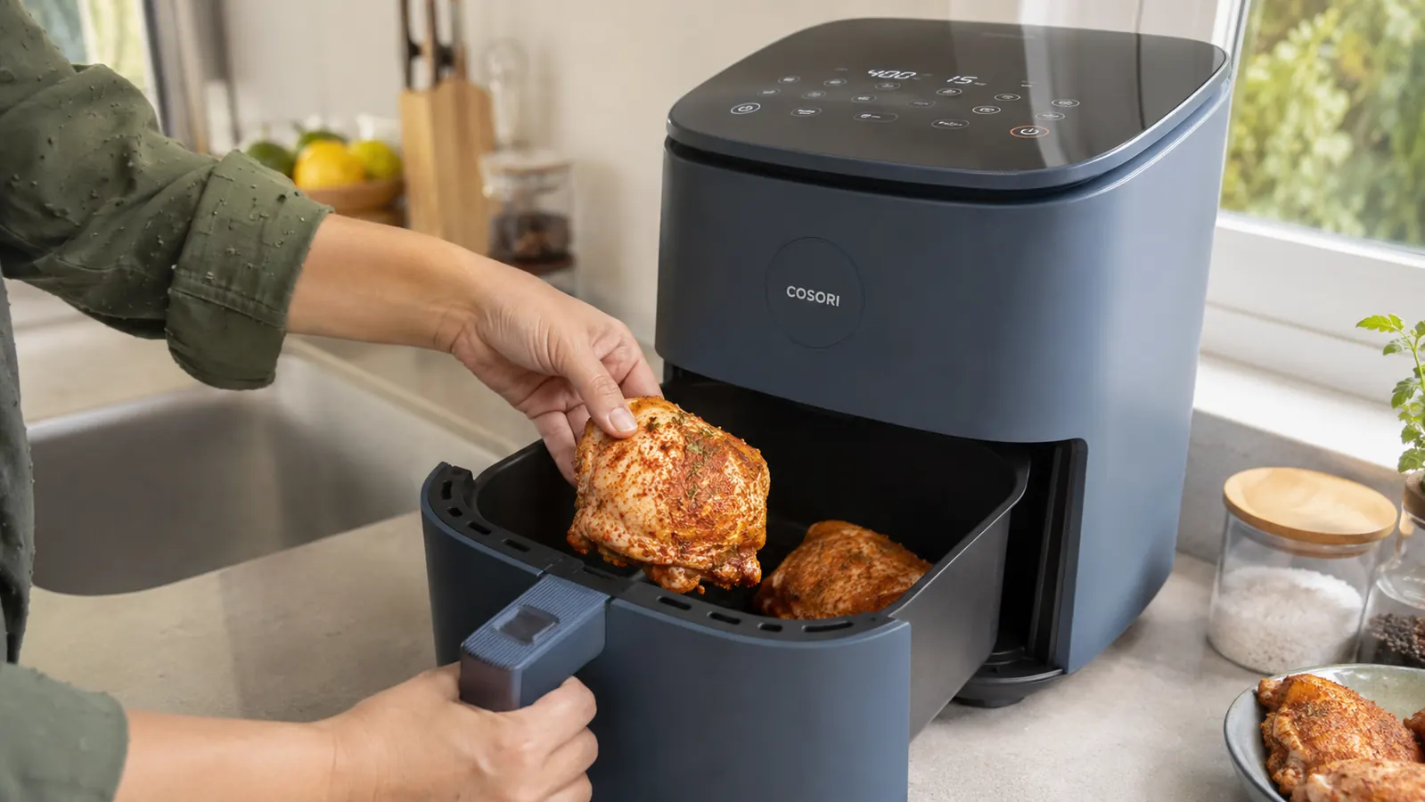 Hand placing seasoned chicken thighs into the basket of a compact air fryer on a small apartment counter