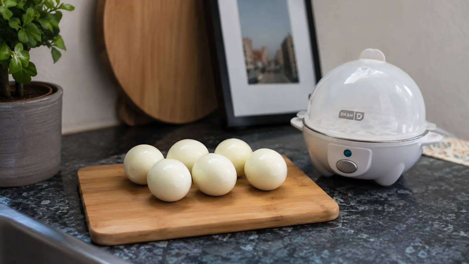 Peeled hard-boiled eggs on a cutting board next to the Dash egg cooker, ready for meal prep