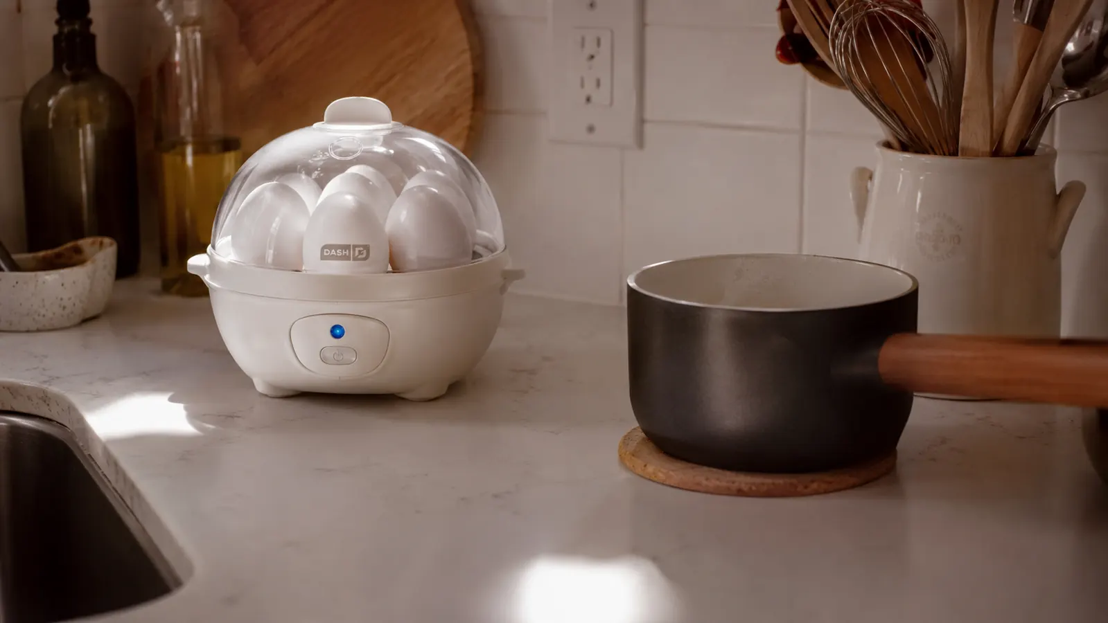 Dash Rapid Egg Cooker sitting on a small apartment kitchen counter next to a pot on the stovetop