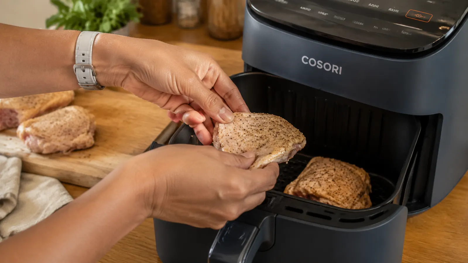 Hand placing seasoned chicken thighs into the Cosori air fryer basket