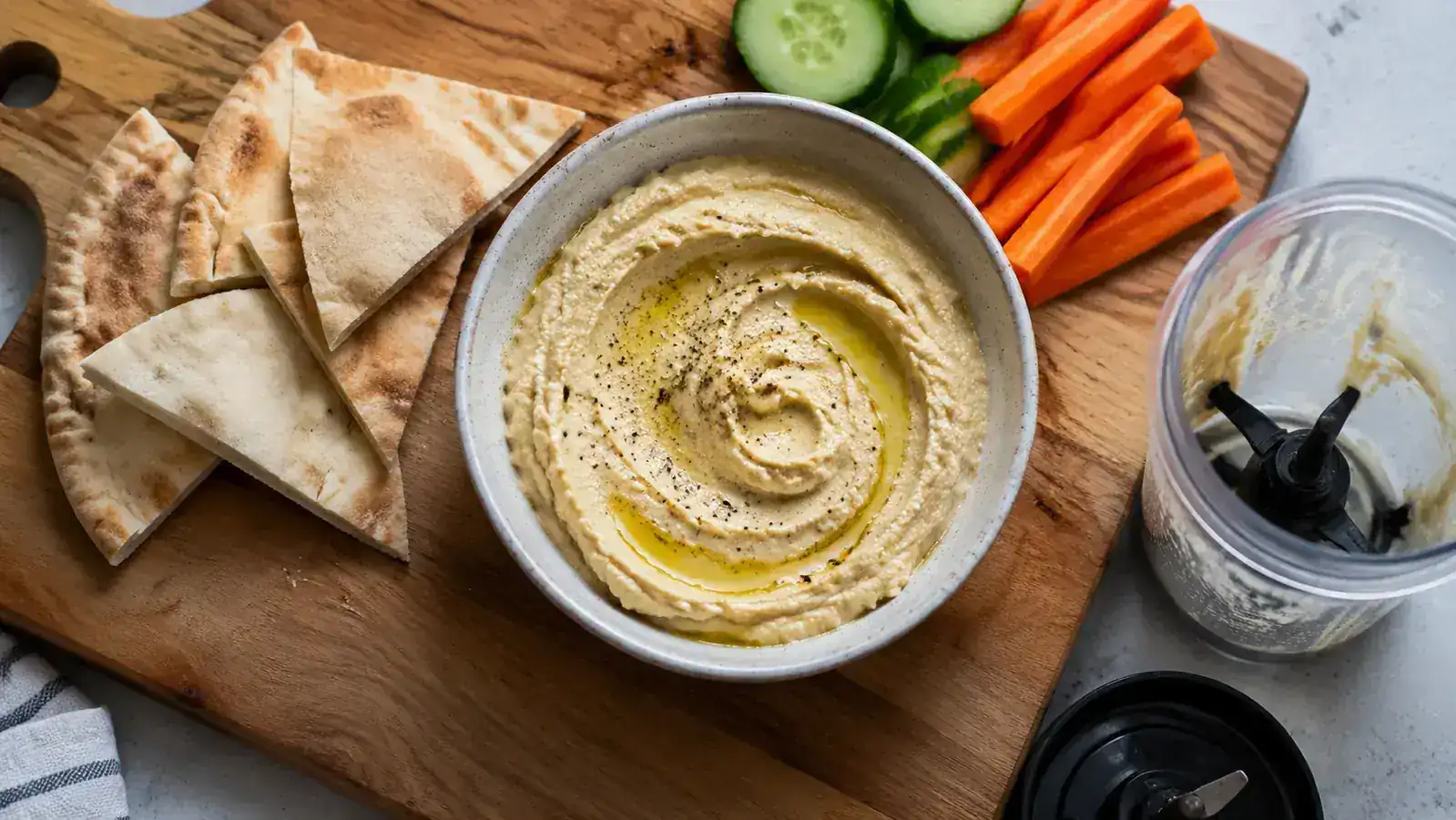 Overhead view of hummus in a small bowl surrounded by pita triangles and chopped vegetables, with the Magic Bullet blender cup to the side