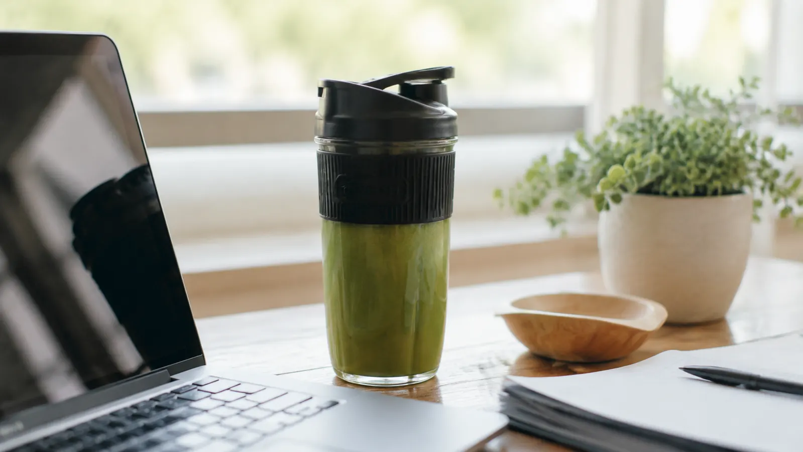 Green smoothie in a small blender cup with a lid, sitting next to a laptop on a small apartment desk, ready to go