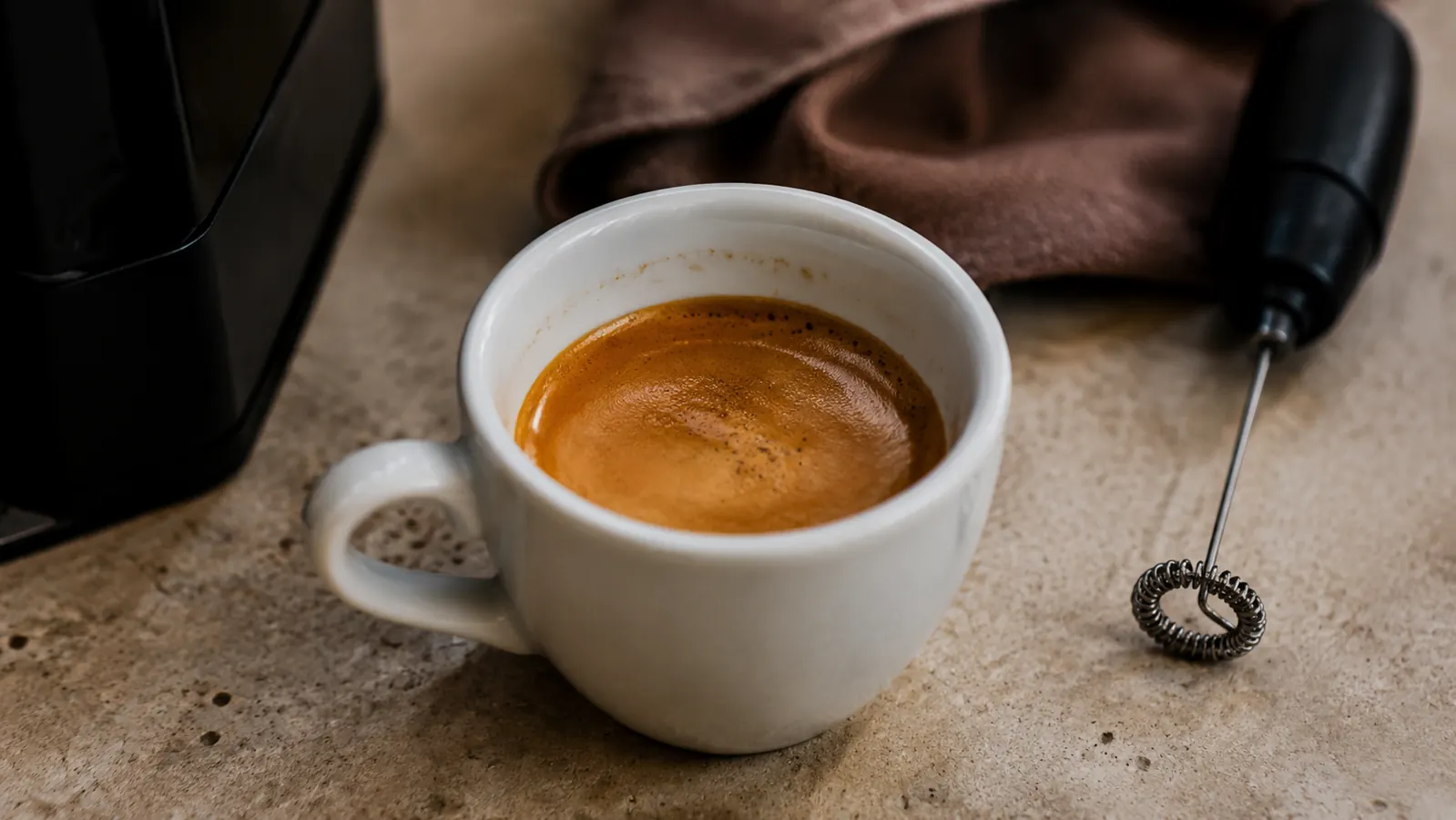 Fresh espresso in a white ceramic cup on a wooden cutting board with a small milk frother beside it on a kitchen counter