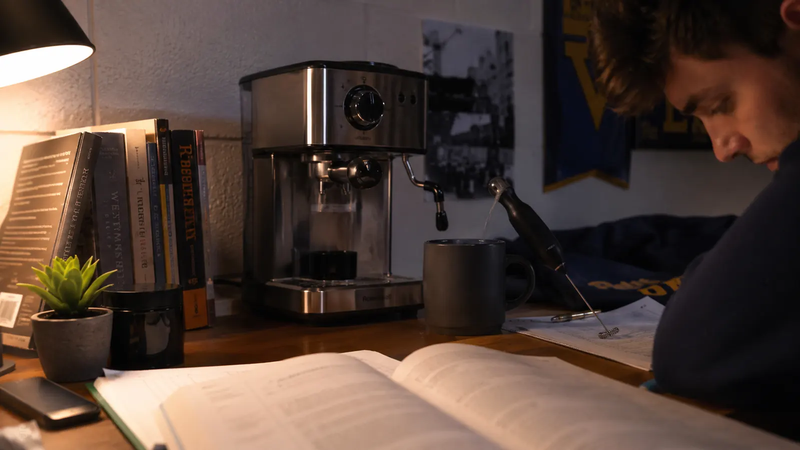 Small espresso machine on a dorm room desk next to a textbook and a small milk frother